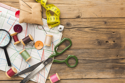 Nähwerkzeuge, grüne Schere, gelbes Maßband, farbige Garnspulen, Nadelkissen, Knöpfe und Schnittmuster auf einem Holztisch